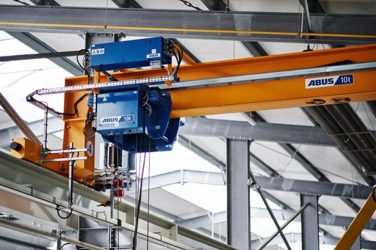 Overhead industrial crane system with a blue ABUS 10-ton hoist mounted on an orange gantry beam inside a manufacturing facility.