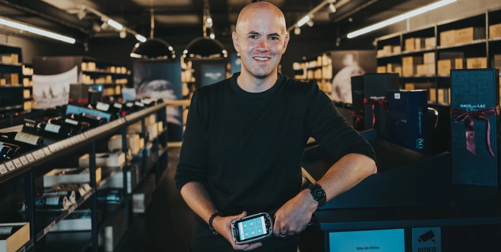 Daniel Hunziker, SAP Administrator at Baur au Lac Vins, holding a mobile scanner device in the warehouse shop area, representing the company's use of SAP-integrated mobile data entry to achieve high inventory accuracy and efficient logistics processes