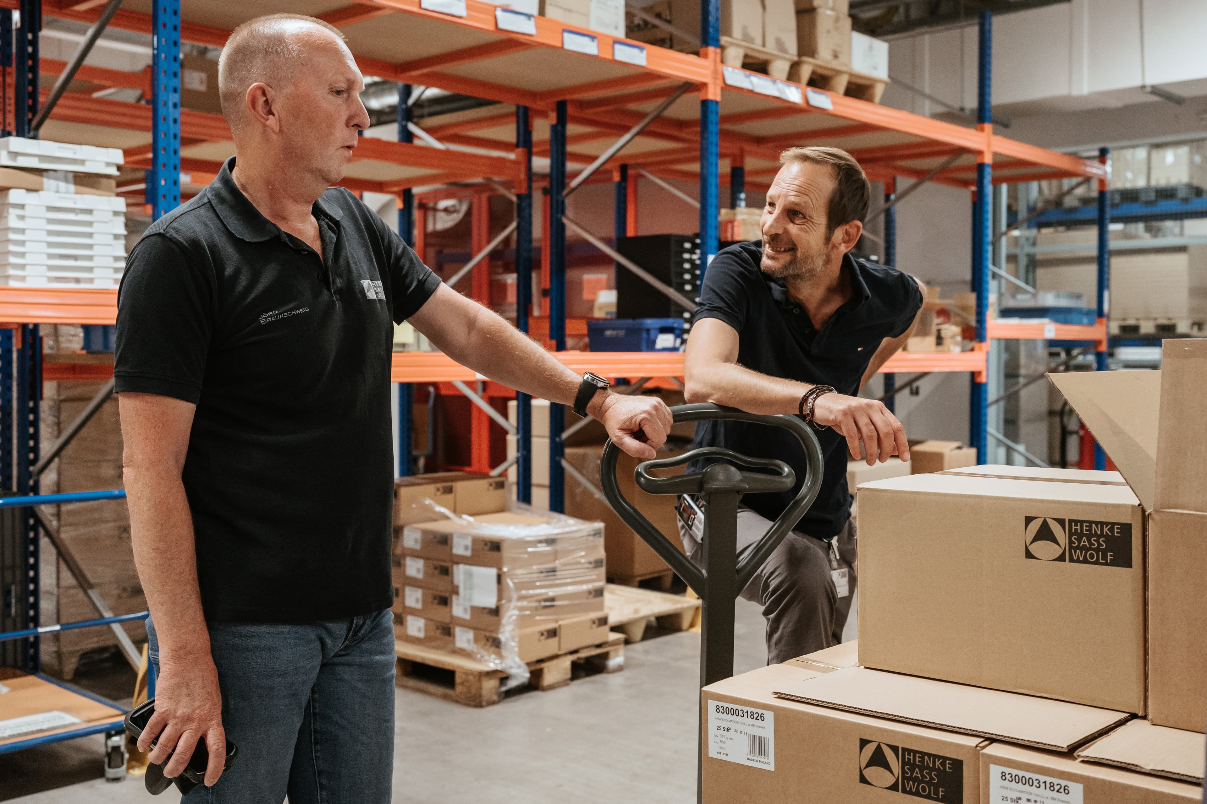 Two Henke-Sass, Wolf warehouse employees discussing operations, one holding a handheld scanner, surrounded by branded shipping boxes and industrial shelving.
