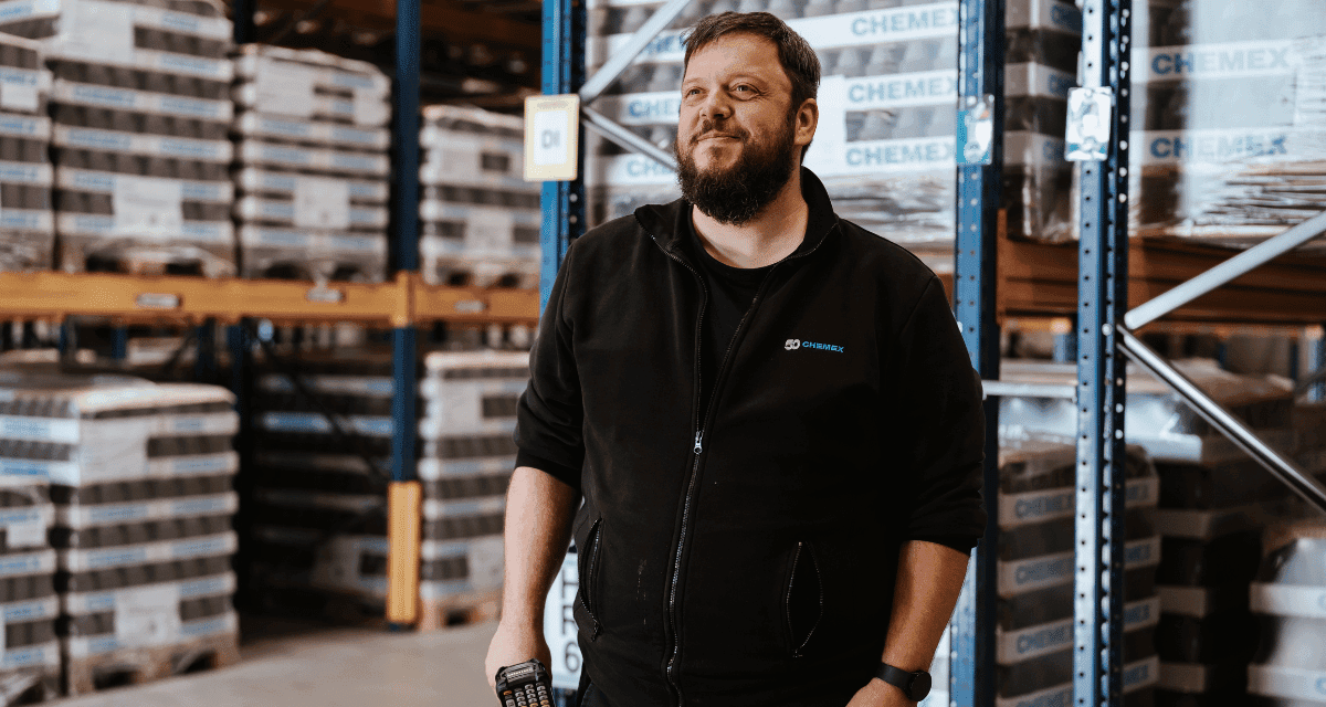 A Chemex warehouse employee holding a mobile barcode scanner, standing in front of stacked pallets of Chemex branded products on industrial shelving units.