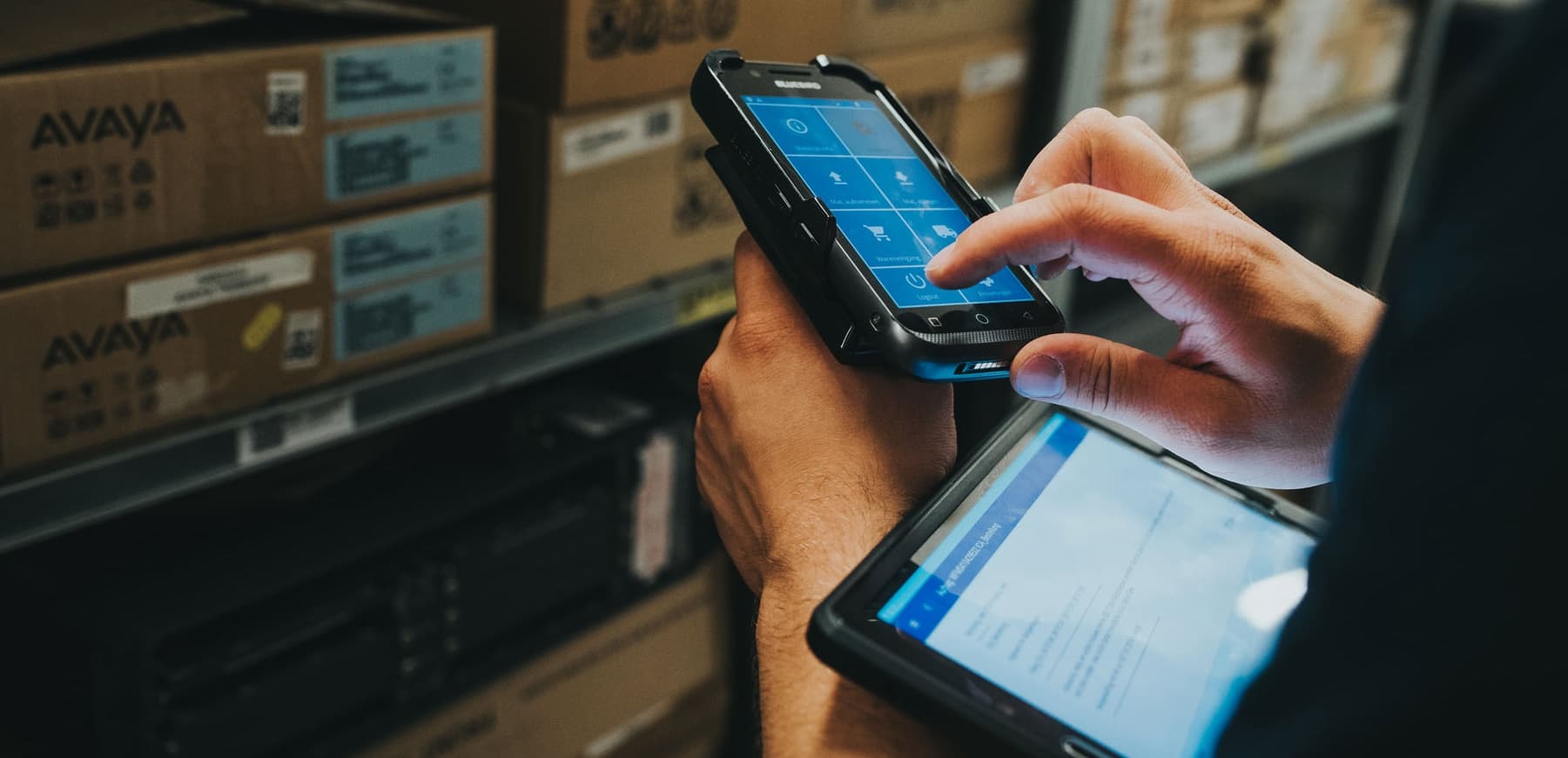A person works in a warehouse using a mobile handheld device with an Ontego screen while also holding a tablet. Stacked boxes on shelves are visible in the background.