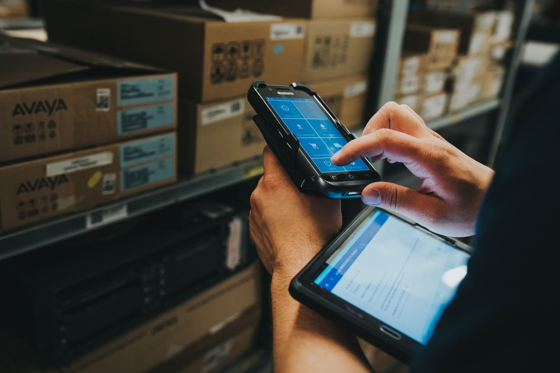 A person works in a warehouse using a mobile handheld device with an Ontego screen while also holding a tablet. Stacked boxes on shelves are visible in the background.