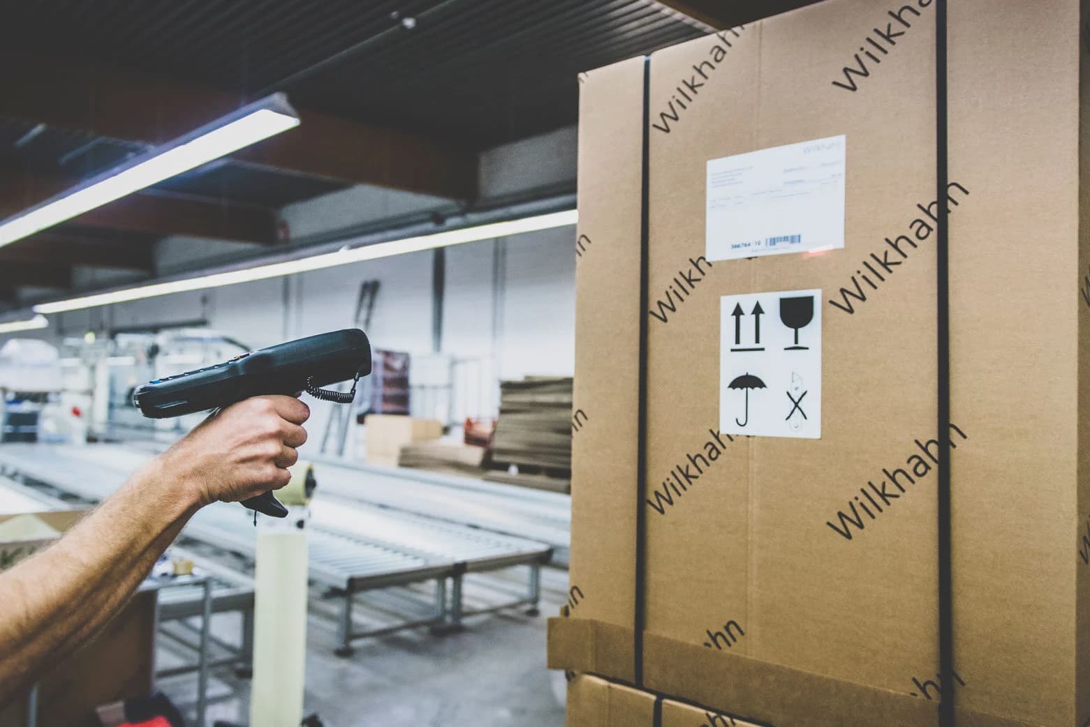 A person's hand holds a barcode scanner, scanning a shipping label on a large cardboard box marked "Wilkhahn" in a warehouse. The box also displays handling symbols for upright, fragile, and keep dry.