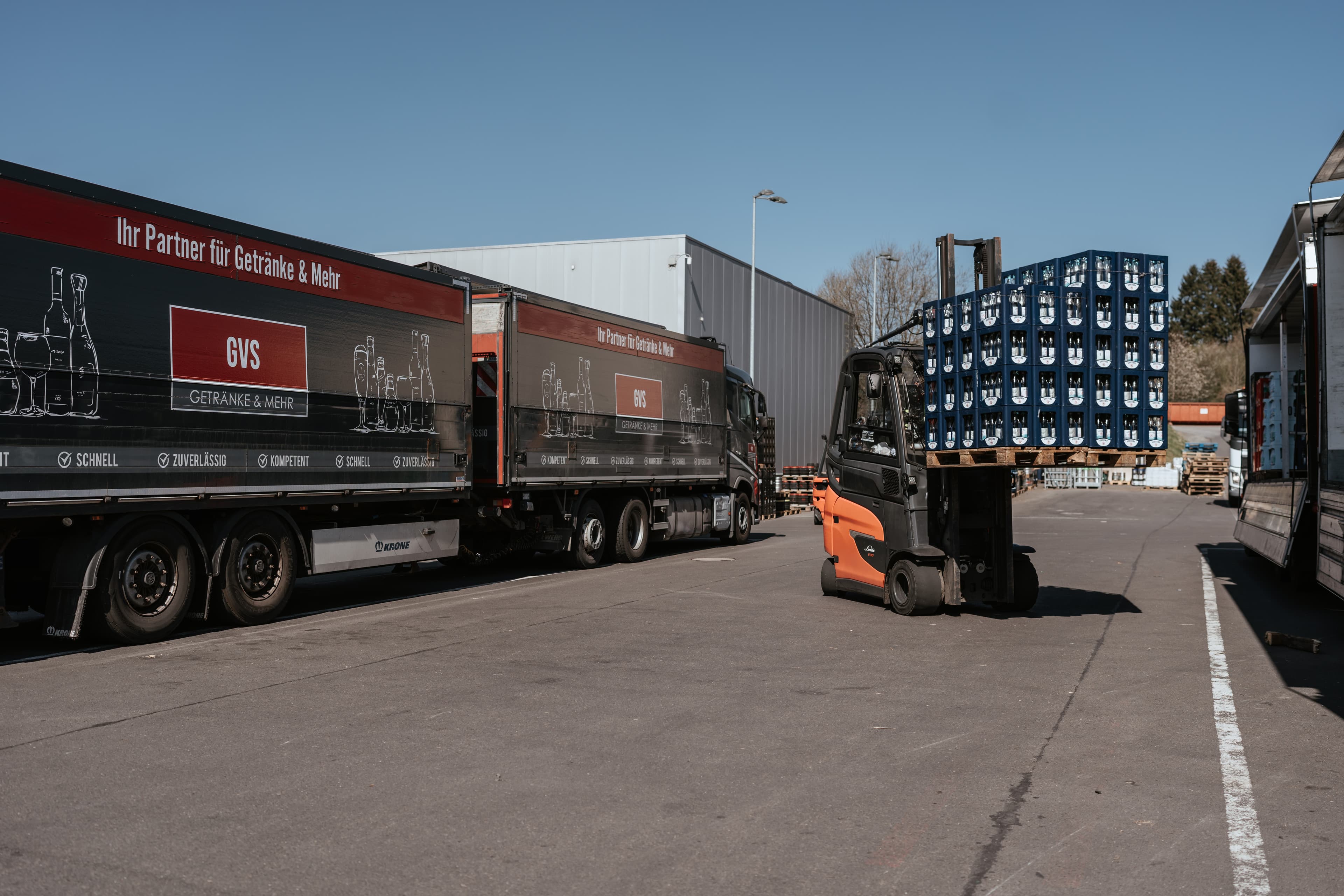 A forklift loading crates of beverages onto a truck at a logistics yard, surrounded by multiple GVS beverage transport trucks